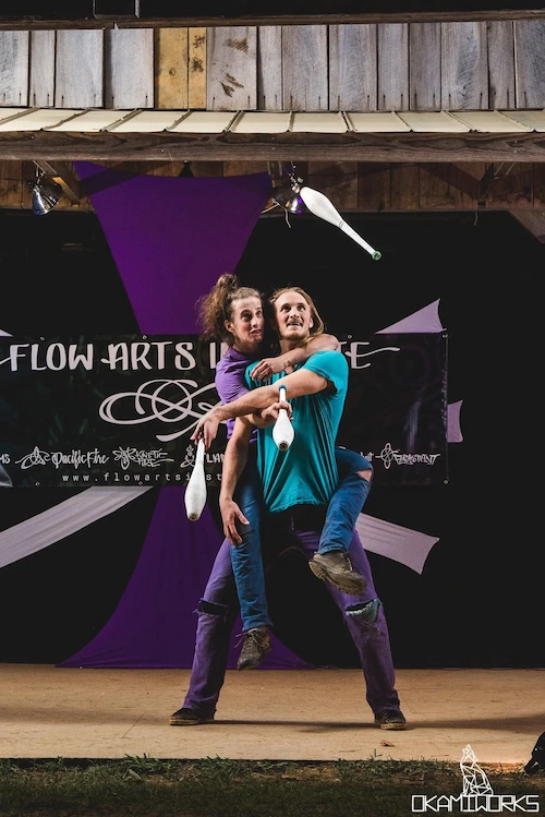 Silly partner jugglers mounted on one another's backs performing at a flow festival