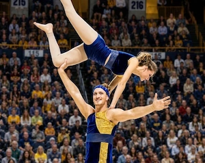 Partner acrobats performing at UTC basketball halftime show 
                    with one holding the other over head by the knee and smiling at audience
