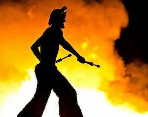 Faun character on stilts dancing with a massive fire in the background